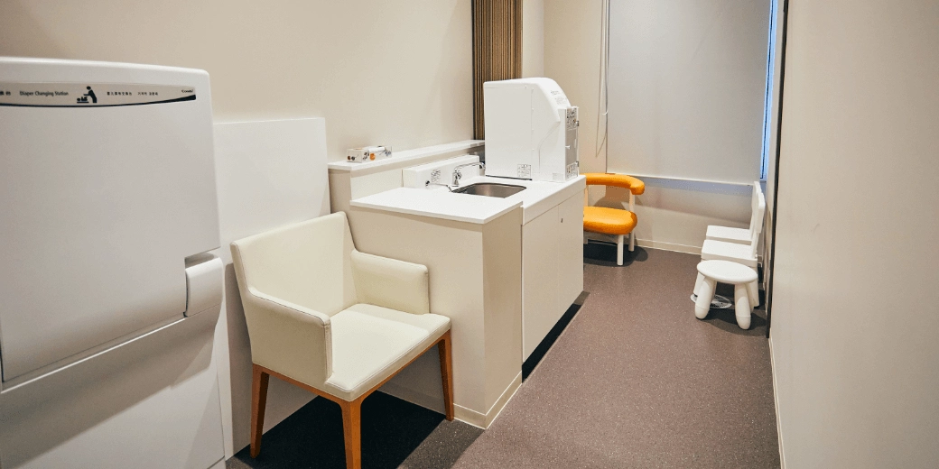 Photo : In the back, there is a window with curtains and four chairs for children, a simple sink in front of it, a white adult chair next to it, and a folding diaper-changing seat in front.