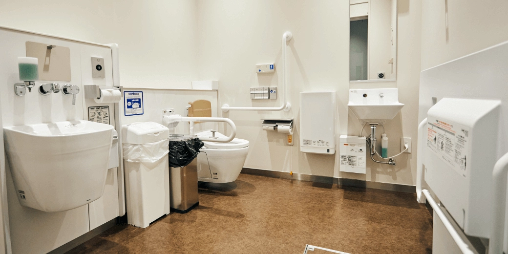 Photo : A toilet with a handrail, a washbasin filled with liquid soap, two types of waste bins (one of which opens and closes with a sensor), facilities for ostomates (persons with artificial anus and artificial bladder) (small in front) with a mirror), there is a folding universal seat