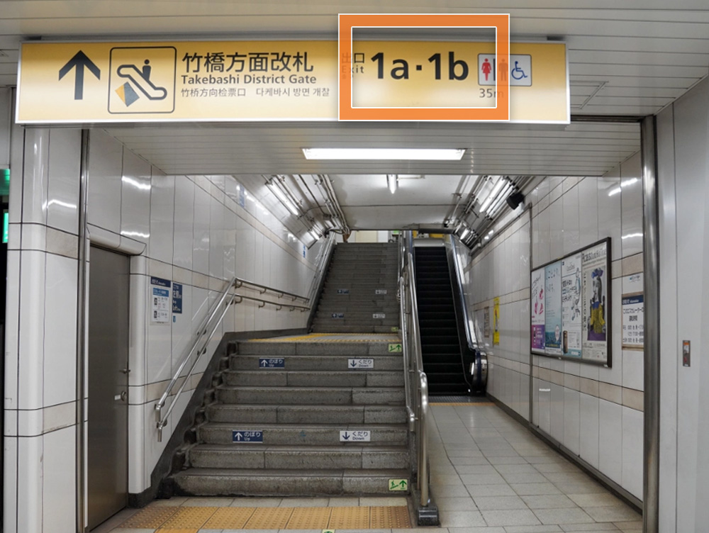 写真：竹橋駅ホーム（案内の看板にエスカレーターマーク、竹橋方面改札 出口 1a・1b、トイレ（男、女、車椅子））