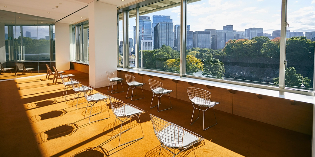 Photo : Eleven chairs are lined up on an orange carpet. The Imperial Palace and the buildings of Otemachi can be seen outside the large windows.