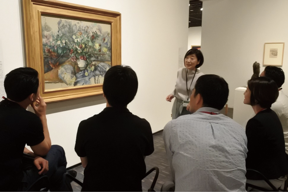 絵画の前で五人の男女が椅子に座って、ガイドスタッフと話しながら鑑賞している写真。