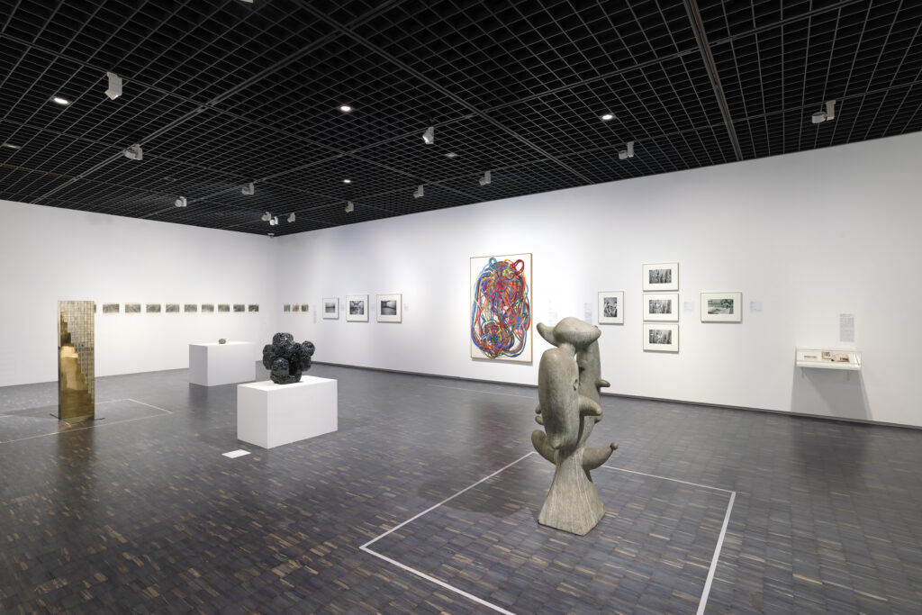 美術館の展示室。中央に彫刻3点。奥の壁には、大きな抽象画油彩と写真作品が複数点展示。
