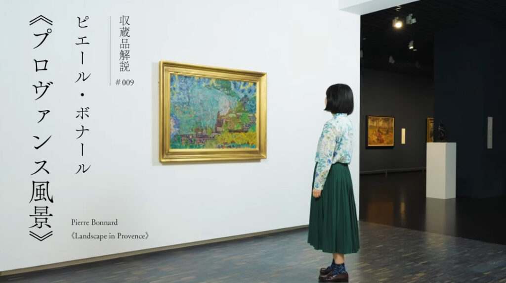 金縁の風景画の前に女性が一人立って、絵を見ている。