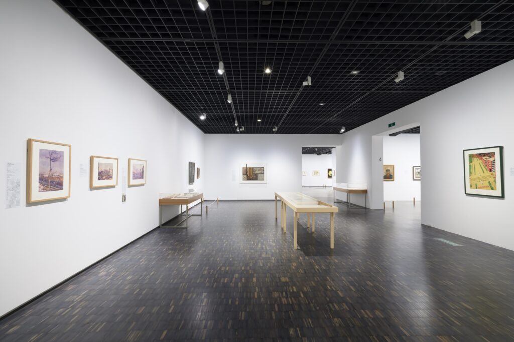 白壁の展示室。壁には水彩画がかけられている。資料を展示する台が3つある。
